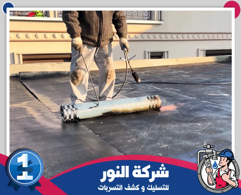 عزل الاسطح من مياه الامطار |شركة النور للتسليك عامل يقوم بفرد وتركيب طبقة عزل على سطح مبنى باستخدام أدوات مخصصة لمنع تسرب المياه، ضمن أعمال شركة عزل اسطح بالنعيرية لحماية الأسطح وتحسين مقاومتها للعوامل الخارجية.