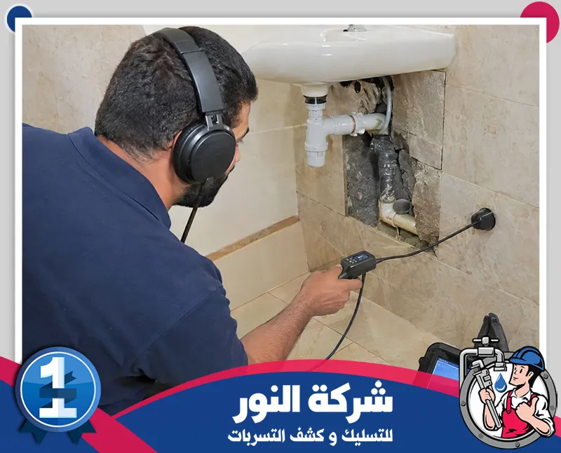 فني يرتدي سماعات ويفحص الجدار بجهاز كشف صوتي بجوار مواسير مكشوفة أسفل المغسلة ومع شعار النور للتسليك