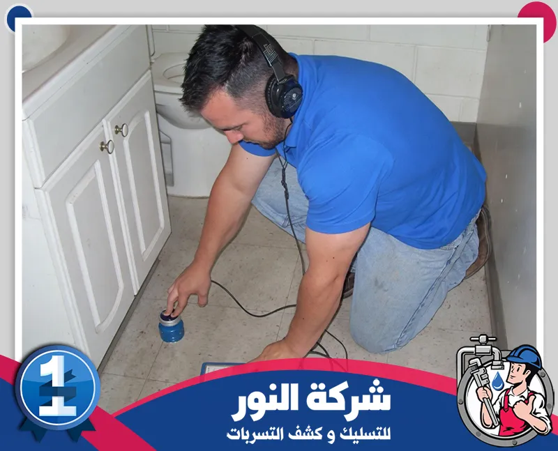 فني بقميص أزرق يرتدي سماعات ويضع حساسًا موصولا بسلك على أرضية الحمام بجوار خزانة بيضاء لفحص التسربات